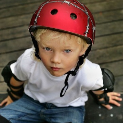 Pojke med röd hjälm, sitter på skateboard och tittar upp i kameran.